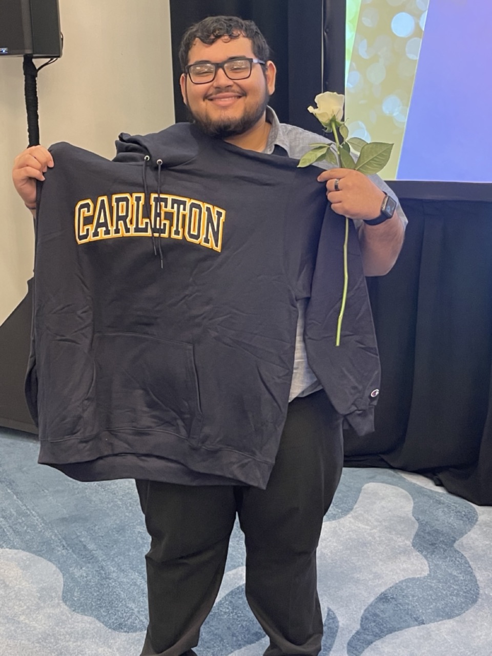 Mmeri scholar smiling as he holds up a Carleton College sweatshirt
